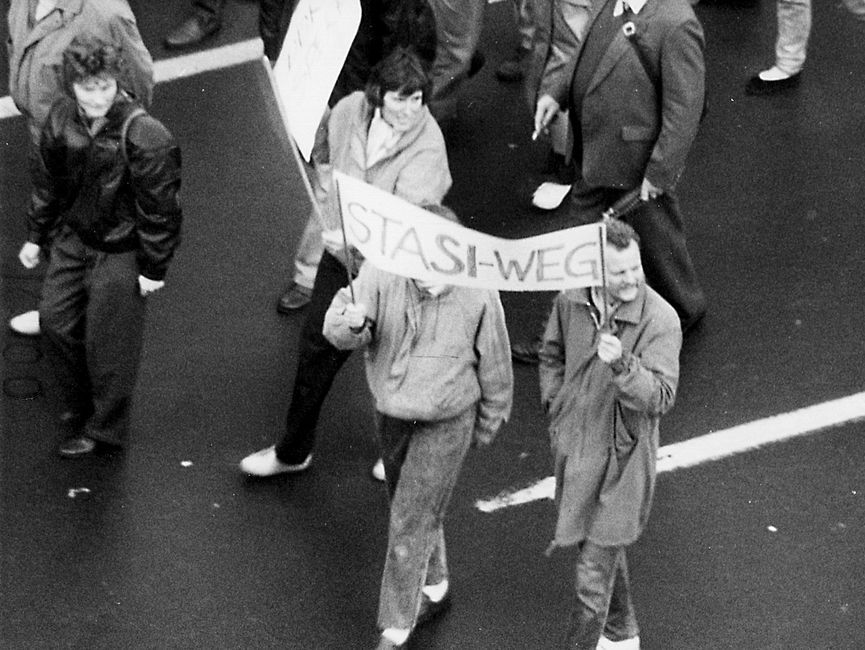 Im Zentrum des Bildes sieht man zwei Personen, die zusammen ein Transparent halten. Darauf steht "Stasi-Weg".