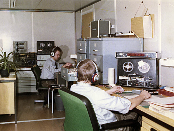 MfS-Mitarbeiter beim Abhören zuvor aufgezeichneter Telefongespräche. Zu sehen sind zwei Männer, die an Tonbandapparaten sitzen und Kopfhörer tragen.