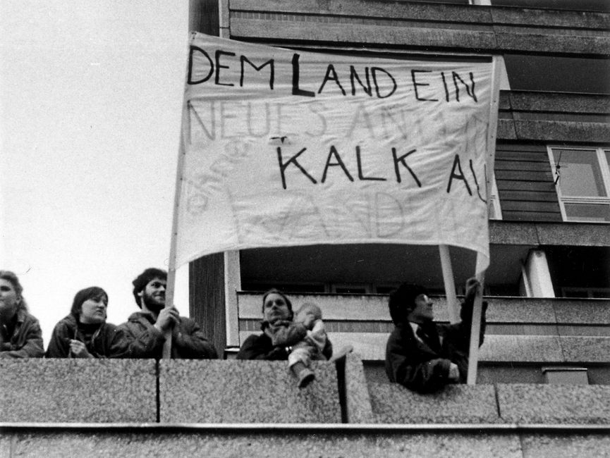 Demonstranten, die auf einem Balkon stehen. Darunter ein Kind auf dem Arm eines Erwachsenen. Zwei Demonstranten halten ein Transparent hoch. Es sind nur die Worte 'Dem Land ein neues...ohne Kalk' zu erkennen.