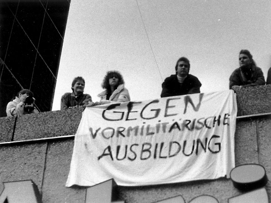 Auf dem Bild sind fünf Personen zu sehen, die auf einem Dach oder Balkon stehen. Ihre Blicke sind nach unten gerichtet. Ein Transparent mit der Aufschrift 'Gegen vormilitärische Ausbildung' ist an der Fassade angebracht. Eine Person fotografiert.