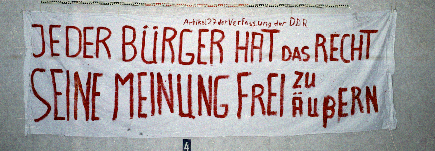 Das Bild zeigt ein abfotografiertes Transparent mit der Aufschrift: ""Artikel 27 der Verfassung der DDR / Jeder Bürger hat das Recht seine Meinung frei zu äußern"