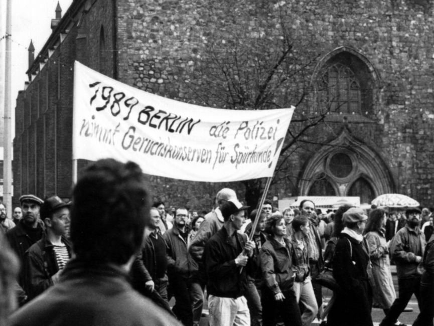 Demonstrationszug, der an einer Kirche vorbeizieht: Zwei junge Männer tragen ein Transparent mit der Aufschrift '1989 Berlin die Polizei nimmt Geruchskonserven für Spürhunde'.