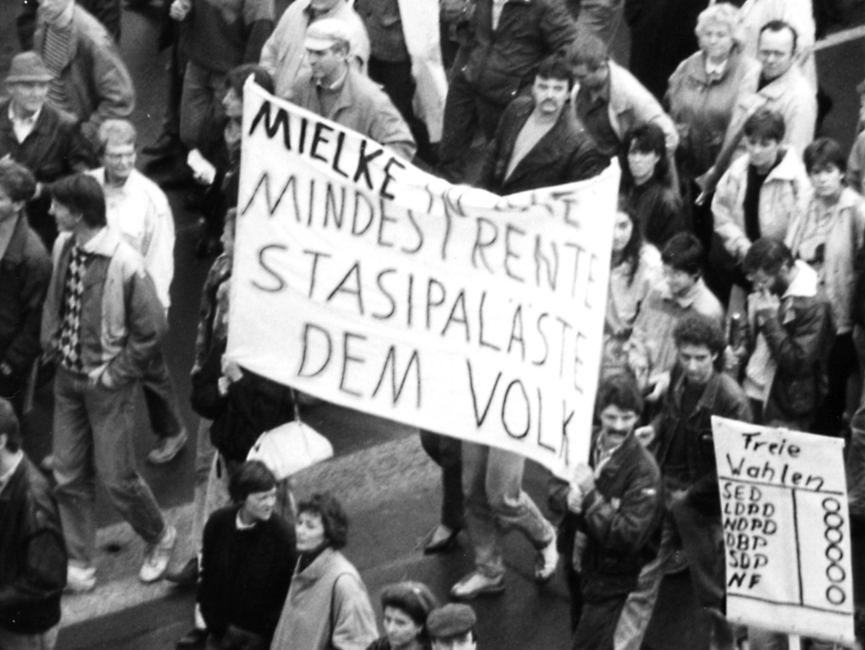 Das Bild zeigt den Blick in einen Demonstrationszug. In der Mitte des Bildes ist ein Transparent mit der Aufschrift 'Mielke [unleserlich] Mindestrente Stasipaläste dem Volk' zu sehen. Unten rechts im Bild ist ein Plakat auf dem 'Freie Wahlen SED LDPD NDPD DBD SDP NF' steht. Hinter den Parteiabkürzungen sind Kreise zum Ankreuzen.