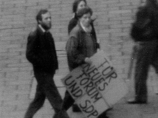 Zu sehen sind drei Männer. Einer von ihnen hält ein Schild mit der Aufschrift 'Für Neues Forum und SDP'