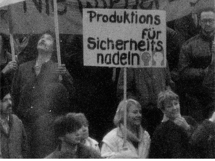Das Bild zeigt einen Blick ganz nah in die Menschenmenge. Eine Frau hält ein Plakat mit der Aufschrift 'Produktions für Sicherheitsnadeln'.