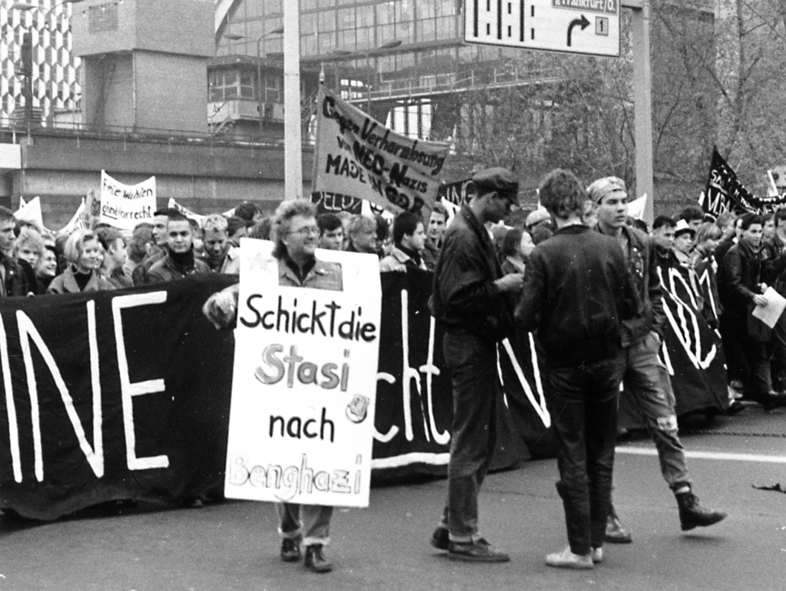 Demonstrierende vor dem S-Bahnhof Alexanderplatz. Im Hintergrund kann man das Centrum Warenhaus erkennen. Einige Demonstranten halten Transparente hoch. Auf einem steht "Freie Wahlen ohne Vorrecht". Es scheint der Anfang des Demonstrationszuges zu sein. Vorne wird ein Banner gehalten. Vor dem Banner steht eine Gruppe aus drei jungen Männern und ein Mann, der ein Plakat trägt mit der Aufschrift "Schickt die Stasi nach Benghazi".