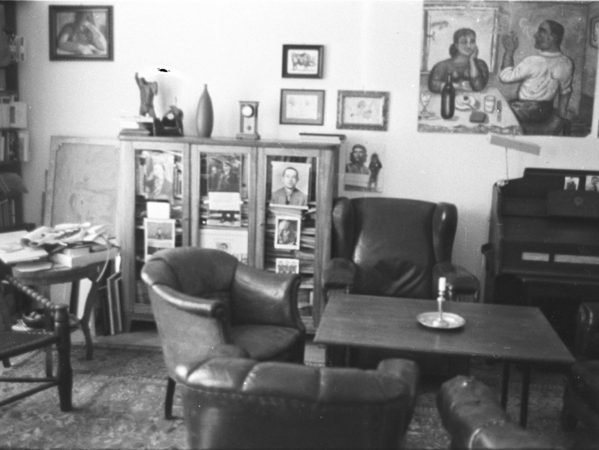 Blick in ein Zimmer, in dem ein Tisch mit Sesseln und ein gefüllter Bücherschrank stehen. An der Wand hängen mehrere Bilder, rechts ist ein verschlossener Sekretär zu sehen.
