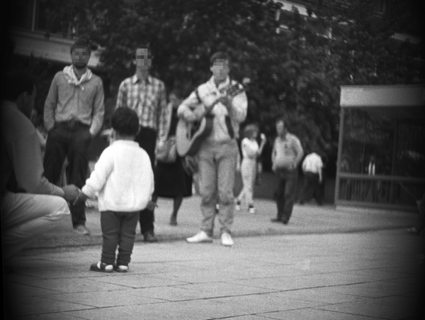 Das Bild zeigt mehrere Menschen auf dem Alexanderplatz in Berlin. Rund um die Aufnahme ist ein kreisrunder, Schatten zu sehen. Vermutlich wurde das Foto aus einer präparierten Tasche oder einem anderen Gegenstand durch ein Loch heraus aufgenommen. Die Perspektive liegt nahezu auf Bodenhöhe. Im Zentrum stehen drei Männer. Einer von ihnen spielt Gitarre. Vor ihnen steht ein Kind. Rund um sie herum sind weitere Personen zu sehen.