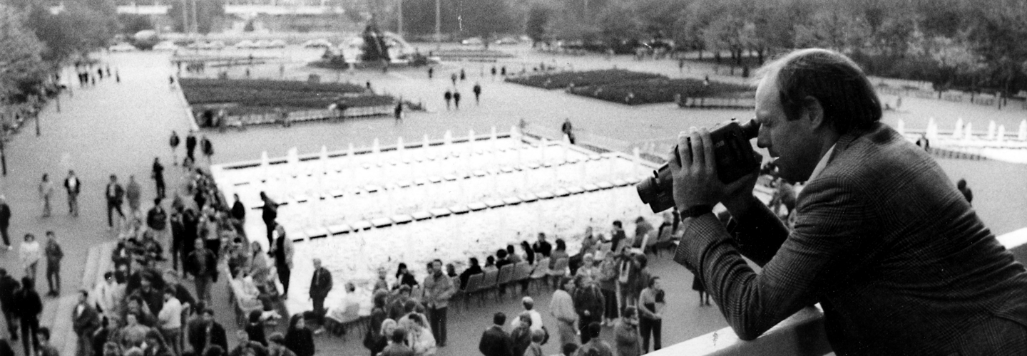 Blick auf den Platz am Neptunbrunnen in Ost-Berlin. Auf dem Platz sieht man eine kleinere Ansammlung von Menschen. Das Bild wurde von einem Balkon aufgenommen. Auf dem Balkon steht ein Mann mit Kamera und filmt das Geschehen auf dem Platz.